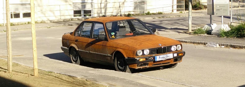 An old BMW with a fully rusted body parked on a street.